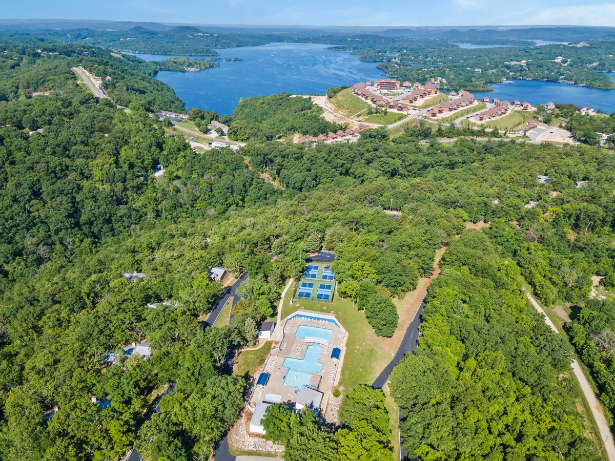 A wide exterior view showing the community swimming pool with a scenic lake in the background, lush greenery surrounding the area, and residential buildings visible at a distance. The image conveys a sense of leisure, recreation, and connection to the waterfront.