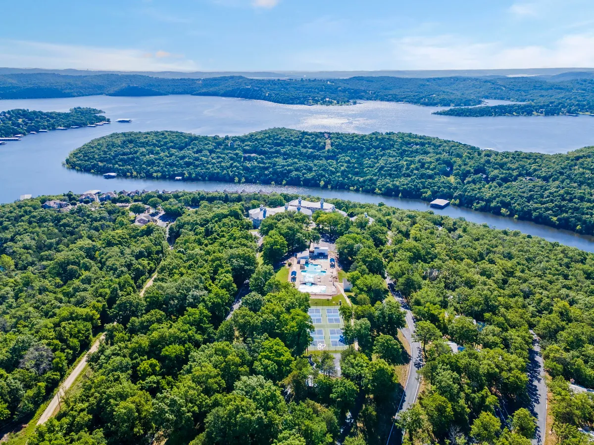 A panoramic exterior scene featuring lush greenery, a broad expanse of Table Rock Lake, scattered residential structures, and recreational facilities. The foreground shows ample open space and gentle shoreline, suggesting easy access to boating and water activities. The peaceful mood is amplified by the sunset hues reflected on the water, hinting at serene evenings and active days outdoors.