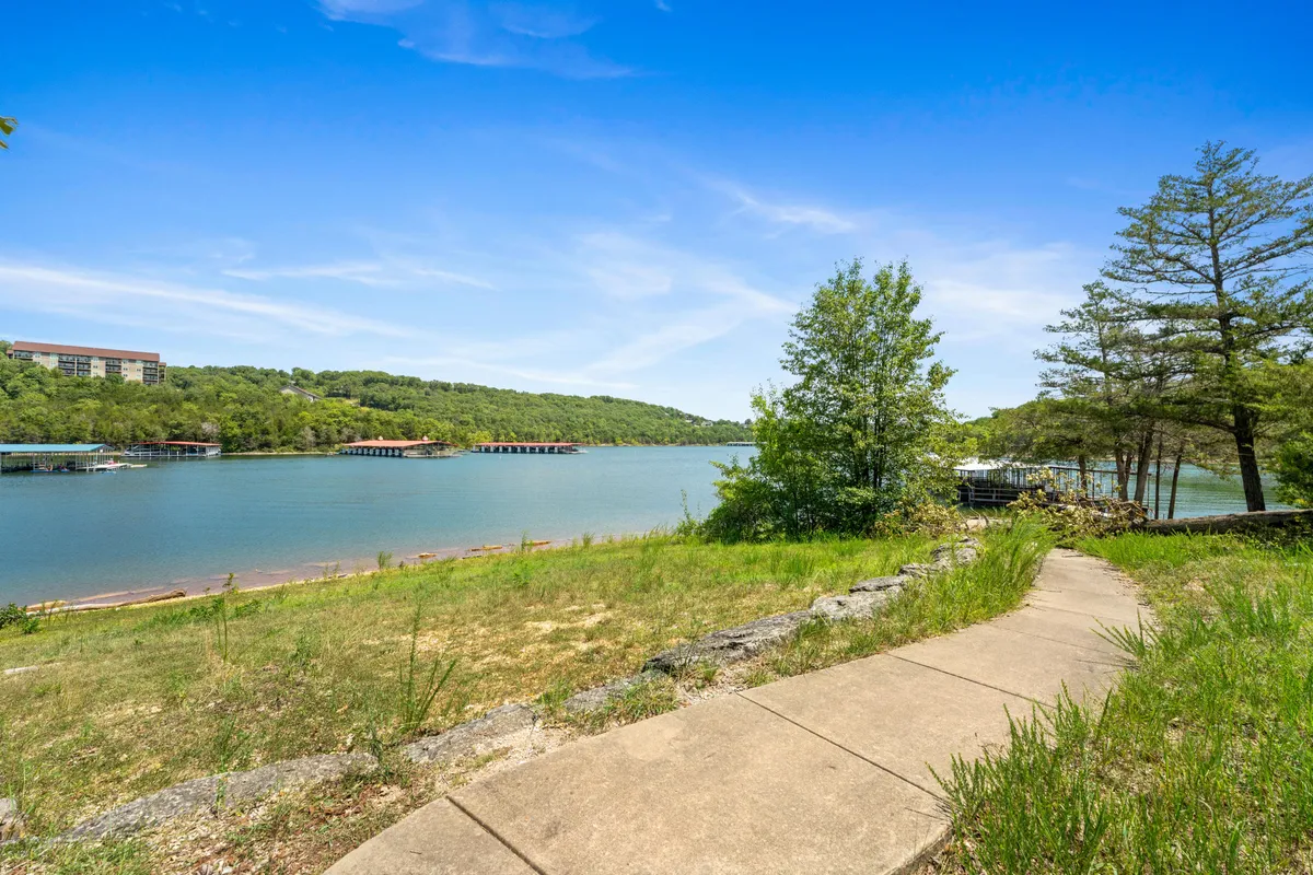 A tranquil view of Table Rock Lake framed by lush greenery and rolling hills, with a pathway leading to a lakeside dock. The serene water reflects the sky, evoking peaceful mornings and opportunities for walking, fishing, or simply relaxing by the shore.