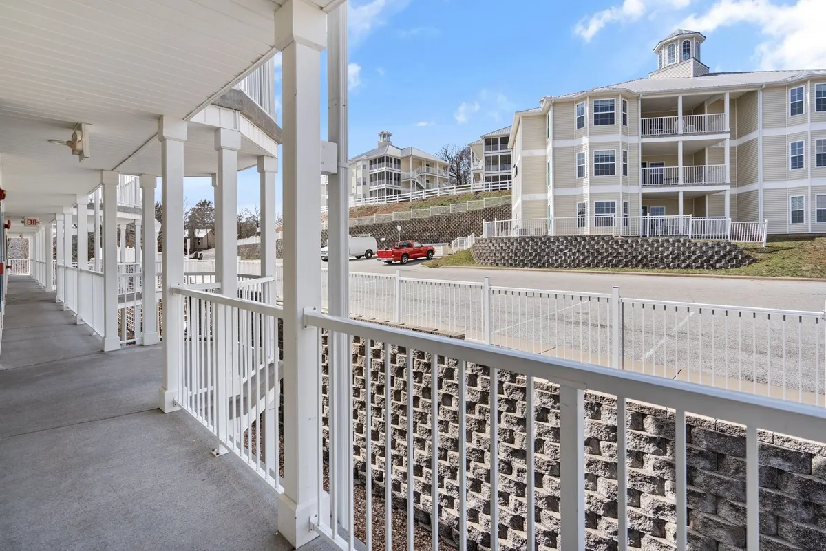This image depicts a private exterior deck or balcony with a white railing, adjacent to a multi-story building. The deck overlooks a peaceful outdoor area, suggesting a spot to enjoy morning coffee or casual gatherings with family and pets. A red truck and stairs are visible, adding a lived-in, approachable feel.