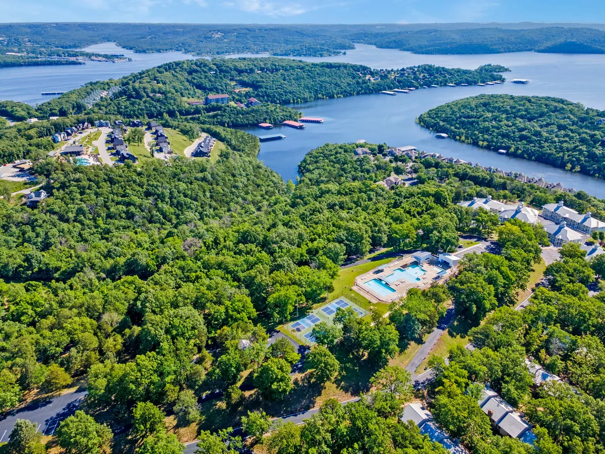 A wide-angle exterior view showing the sparkling lake, swimming pool, tennis courts, and residential buildings, all surrounded by lush green hills. The scene is calm yet inviting, suggesting a range of on-site activities from swimming to tennis to lakeside relaxation.