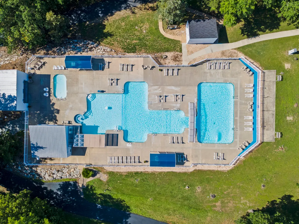 A vibrant exterior view of the resort's large swimming pool, flanked by smaller pools and surrounded by sun loungers set on lush green grass. The scene shows guests swimming, sunbathing, and socializing, capturing the leisurely and refreshing atmosphere of the pool area during peak season.