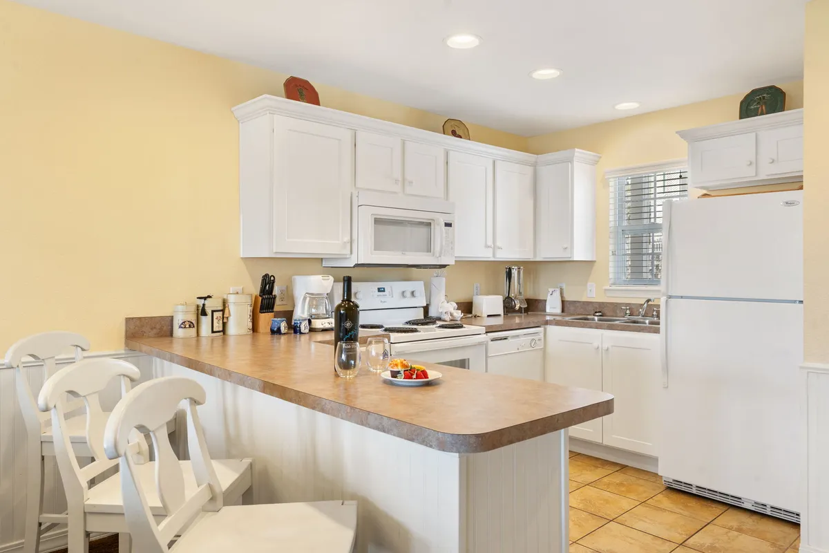 The image showcases a bright, inviting kitchen with a spacious island at the heart, gleaming white cabinetry, and thoughtful touches like a wine bottle, glasses, and fresh fruit on the countertop. The space radiates warmth and serves as a perfect gathering spot for families or friends to cook, share meals, and create lasting memories.