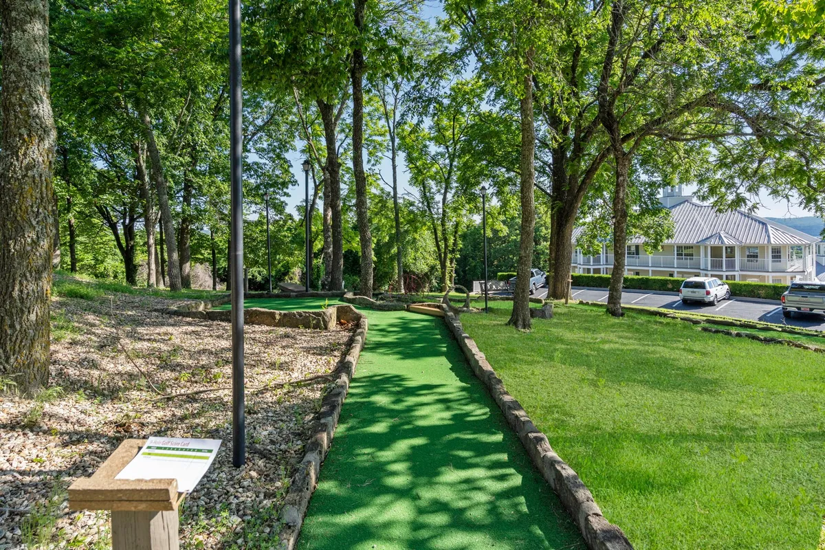 A playful scene featuring a mini-golf pathway winding beneath tall trees, with lush greenery and an informational sign visible. The area appears inviting for families, with space for both mini-golf and nearby picnics or gathering spots.