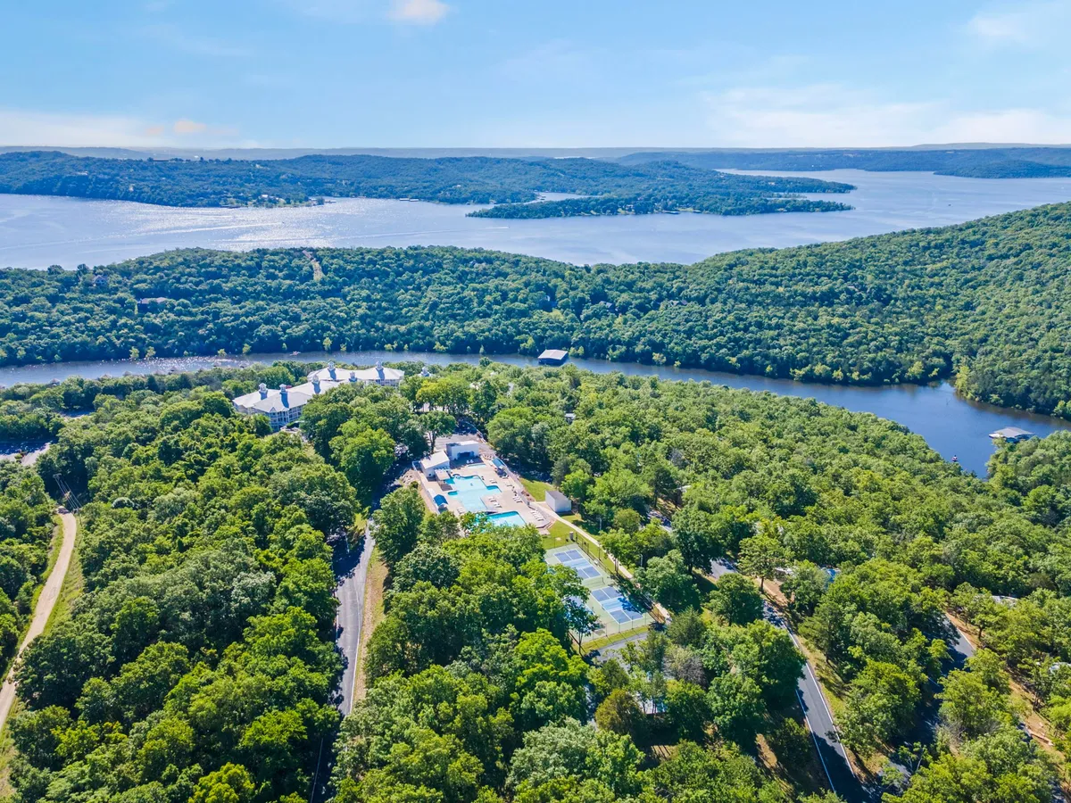 An elevated perspective featuring lush green trees, the sparkling lake, the resort’s swimming pool, and tennis courts. The view captures the inviting outdoor environment, perfect for exploring, swimming, or picnicking by the water's edge.