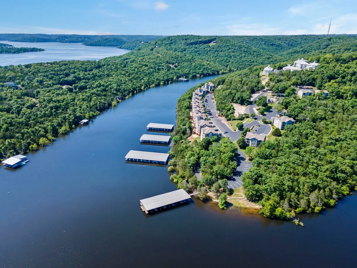 A tranquil, wide-angle exterior of Table Rock Lake with docks, a marina, lush surrounding greenery, and residential buildings in the background. Boaters, anglers, and hikers are suggested by the layout, and the calm waters evoke moments of relaxation or adventurous water sports.